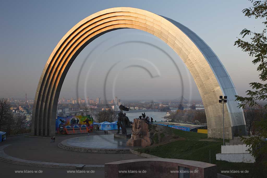 Kiew Blick vom Vladimirskaya Gorka Park durch den Bogen der Voelkerfreundschaft zwischen der Ukraine und Russland auf den Fluss Dnepr sowie die Hochhaeuser  am Westufer im roetlichen Licht der untergehenden Sonne . The friendship Arch: sculptor A. Skoblikov and Architect I. Ivanov and others. Dedicated to the unification of Russia and Ukraine, but called "The Yoke" by kievans. The rainbow shape arch is 50 metres in diameter. Viewing deck were most of the east bank can be viewed, Troeschina and towards the north of city. Podil and Obolon. Undemeath the Friendship Arch are two statues. The left one is made from bronze and depicts a Russian and Ukrainian worker holding up the Soviet Order of Friendship of People the other one is made from granite and depicts the particants of the Pereyaslavska Rada of 1654.