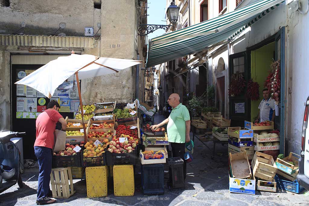 Blick in eine Gasse in der Stadt Pizzo in Kalabrien, Vibo Valentia, Italien mit Sicht auf ein Alimentari, Lebensmittelgeschaeft mit Obst und Gemueseauslage auf der Strasse, dabei der Haendler und ein Kunde